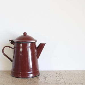 May include: A maroon enamel coffee pot with a lid and handle. The pot has a spout for pouring and a white rim around the lid. The pot is on a light-coloured surface against a white background.