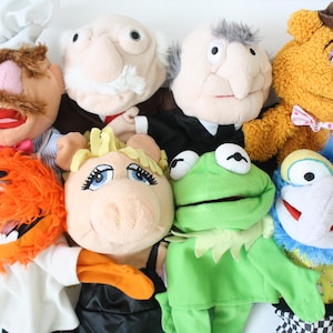 Op de afbeelding: Een verzameling van acht Muppet-handpoppen, waaronder een kok, twee oudere mannen, een bruine beer met een vlinderdas, een oranje monster, een varken, een groene kikker en een blauw wezen met een grote neus, allemaal samen in een groep.