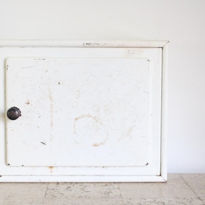 Peut inclure: Une armoire murale rectangulaire en métal blanc dotée d'une porte battante unique avec un petit bouton rond foncé. La surface de l'armoire présente des signes d'usure visibles, notamment des rayures légères, des éraflures et des taches de rouille.