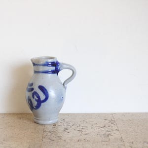 May include: A stoneware pitcher with a light gray base and blue accents. The pitcher features a handle and is decorated with blue swirl designs and bands around the neck. The pitcher is a classic piece of pottery.