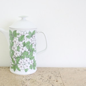 Könnte beinhalten: Weißer Keramik-Krug mit einem Blumendesign mit weißen Blüten und grünen Blättern. Der Krug hat einen Deckel und einen Henkel.