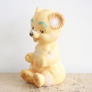 May include: A vintage yellow rubber toy bear with painted eyes and a black nose. The bear is sitting with its legs crossed and its head tilted to the side.