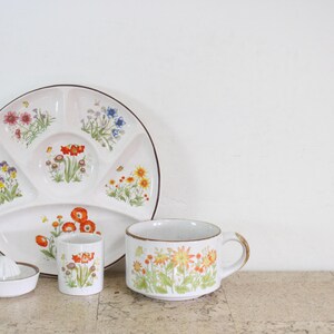 May include: A white ceramic divided plate, a small juice glass, a citrus reamer, and a mug, all decorated with colourful floral patterns featuring red, orange, yellow, blue, and purple wildflowers on a white background with brown rim accents.