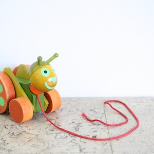 Puede incluir: Un juguete de madera verde y naranja con forma de insecto con una cara sonriente y grandes lunares verdes en las ruedas. El juguete está unido a una cuerda roja.