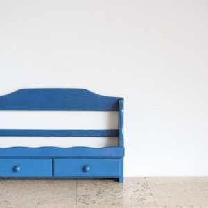 May include: A blue wooden wall shelf featuring an open top section with two horizontal slats and a base section containing two small drawers, each equipped with a single round knob. The unit has a curved top edge and a scalloped trim detail.