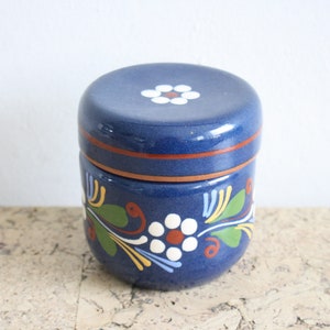 May include: A blue ceramic jar with a lid, decorated with white flowers and green leaves. The jar has a brown rim and a white flower on the lid.