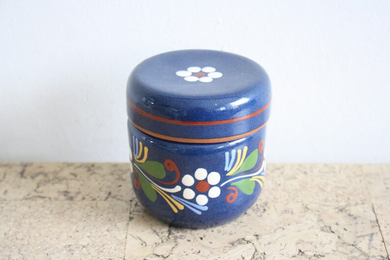 May include: A blue ceramic jar with a lid, decorated with white flowers and green leaves. The jar has a brown rim and a white flower on the lid.