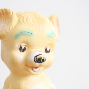 May include: A vintage yellow rubber toy bear with blue painted eyes and a black nose. The bear has a wide open mouth with a pink tongue.
