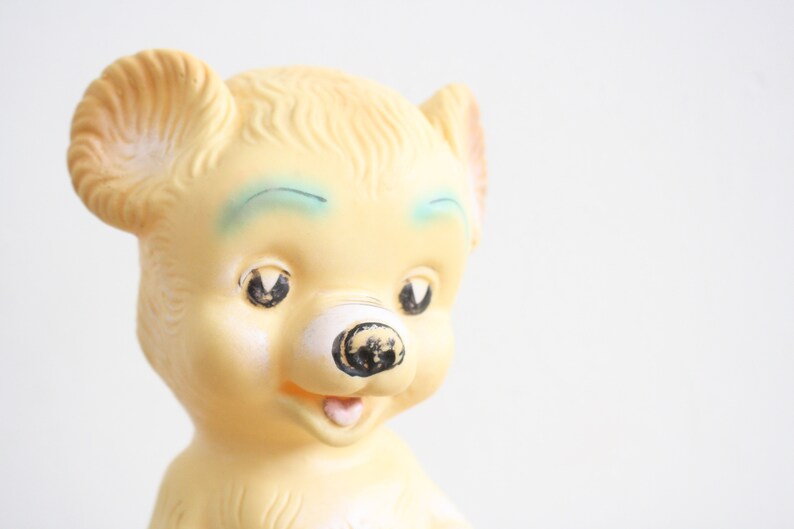 May include: A vintage yellow rubber toy bear with blue painted eyes and a black nose. The bear has a wide open mouth with a pink tongue.