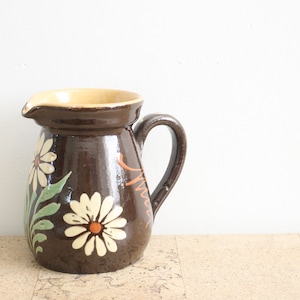 May include: A brown ceramic pitcher with a yellow rim and a floral design featuring white daisies with orange centers and green leaves.