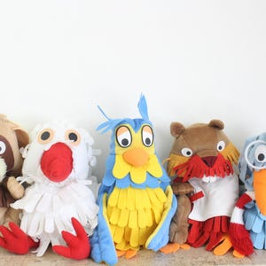 May include: A collection of five handcrafted felt animal puppets featuring a tan raccoon, a white bird with red feet, a blue and yellow owl, a brown beaver with a red mustache, and a blue bird wearing a black cap, glasses, and a red and white scarf.