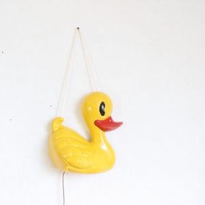 May include: A yellow duck-shaped wall decoration with a red beak and black eyes. The duck is hanging from a white string against a white wall. The duck is a vintage or retro style.