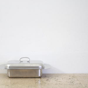Puede incluir: Un recipiente rectangular de acero inoxidable para alimentos con tapa y asa. El recipiente est&aacute; sobre una superficie de color claro contra una pared blanca. La tapa tiene un asa curvada.