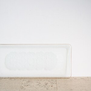 Könnte beinhalten: Ein rechteckiges, klares Glastablett mit einem dezenten, strukturierten Muster in der Mitte. Das Tablett hat einen dünnen, goldfarbenen Rand und steht auf einer hellen, strukturierten Oberfläche vor einer weißen Wand.