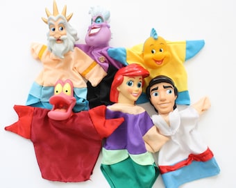 May include: A collection of hand puppets featuring characters from Disney's The Little Mermaid. The puppets include King Triton, Ursula, Sebastian, Ariel, Prince Eric, and Flounder. Each puppet has a fabric body and a plastic head.