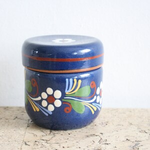 May include: A blue ceramic jar with a lid, decorated with white flowers and green leaves. The jar has a brown rim.