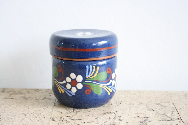 May include: A blue ceramic jar with a lid, decorated with white flowers and green leaves. The jar has a brown rim.