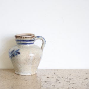 May include: A vintage ceramic pitcher with a handle, featuring a speckled beige body and a brown rim. Blue stripes and a floral design adorn the pitcher. The pitcher is displayed on a light-colored surface against a white background.