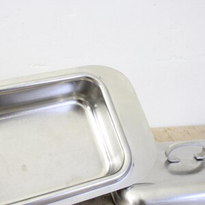 Puede incluir: Una fuente rectangular de acero inoxidable con tapa a juego. La fuente est&aacute; abierta, revelando su interior. La tapa est&aacute; al lado de la fuente, con un asa curvada. Los art&iacute;culos est&aacute;n sobre una superficie de color claro.