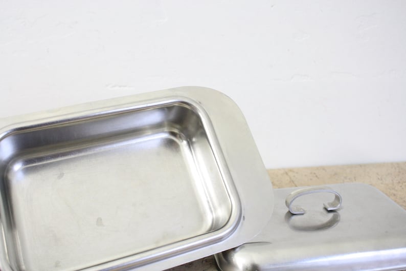 Puede incluir: Una fuente rectangular de acero inoxidable con tapa a juego. La fuente est&aacute; abierta, revelando su interior. La tapa est&aacute; al lado de la fuente, con un asa curvada. Los art&iacute;culos est&aacute;n sobre una superficie de color claro.