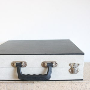 Könnte beinhalten: Ein weißer und schwarzer Vintage-Koffer mit einem schwarzen Griff und silbernen Verschlüssen.
