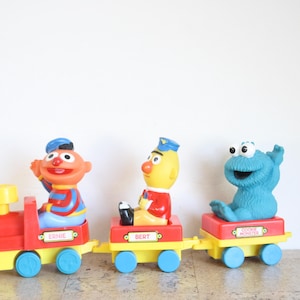 Puede incluir: Un tren de juguete colorido con los personajes de Barrio Sésamo, Epi, Blas y Monstruo de las Galletas. El tren es rojo, amarillo y azul, con cada vagón etiquetado con el nombre del personaje. Un juguete coleccionable divertido.