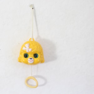 May include: A yellow plastic baby mobile shaped like a puppy's head, with a white bow and green eyes. The mobile hangs from a white string and has a yellow ring at the bottom. The background is a plain white wall.