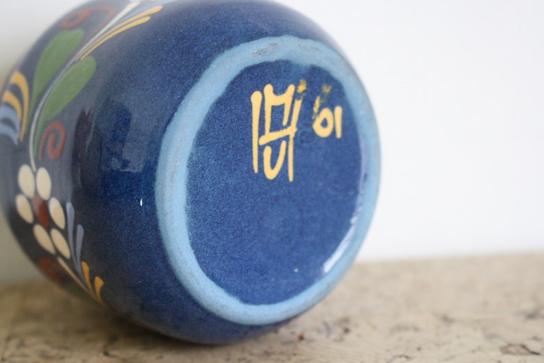 May include: Blue ceramic vase with a white and yellow floral design. The bottom of the vase has a light blue ring and the number "01" written in yellow.
