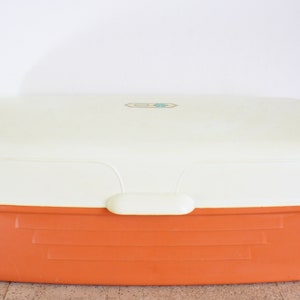 Può includere: Un porta pane in plastica bianco e arancione con un coperchio. Il coperchio ha un piccolo adesivo blu chiaro con un disegno.