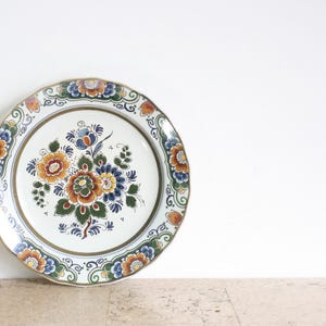 Può includere: Un piatto in ceramica bianca con un motivo floreale blu, bianco e arancione. Il piatto ha un bordo dorato ed è decorato con un motivo tradizionale olandese di Delft.