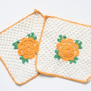 Puede incluir: Dos agarraderas de olla de crochet blanco con diseños florales naranjas y verdes.