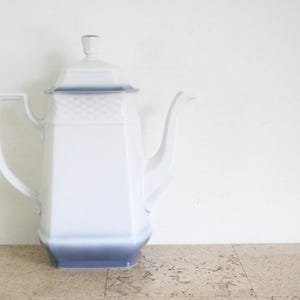 May include: A white porcelain coffee pot with a square base and body, featuring a subtle blue gradient at the bottom and top rim. The pot includes a matching lid with a small knob, a curved handle, and a spout, with a textured basket-weave pattern.