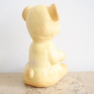 May include: A yellow rubber toy bear sitting on a cork surface. The bear has a textured surface and is in a sitting position with its head turned to the side.