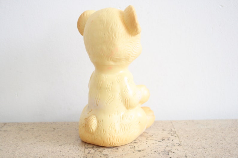 May include: A yellow rubber toy bear sitting on a cork surface. The bear has a textured surface and is in a sitting position with its head turned to the side.