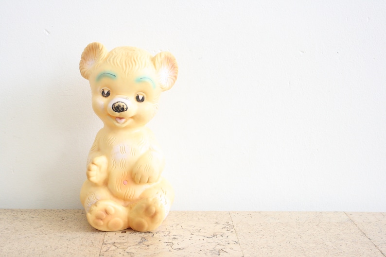 May include: A yellow rubber toy bear with a painted face and black eyes. The bear is sitting with its legs crossed and its arms at its sides.