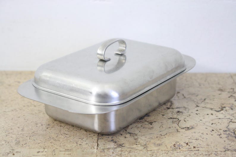 Puede incluir: Una fuente rectangular de acero inoxidable con tapa y asa. La fuente tiene una forma ligeramente redondeada y se encuentra sobre una superficie de color claro. La tapa presenta un asa curvada.