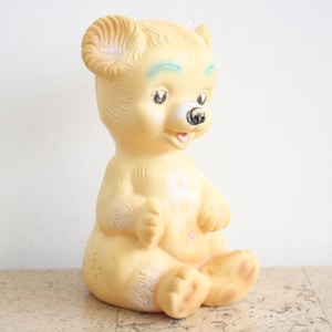 May include: A vintage yellow rubber toy bear with blue eyes and a black nose. The bear is sitting with its legs crossed and has a small smile.