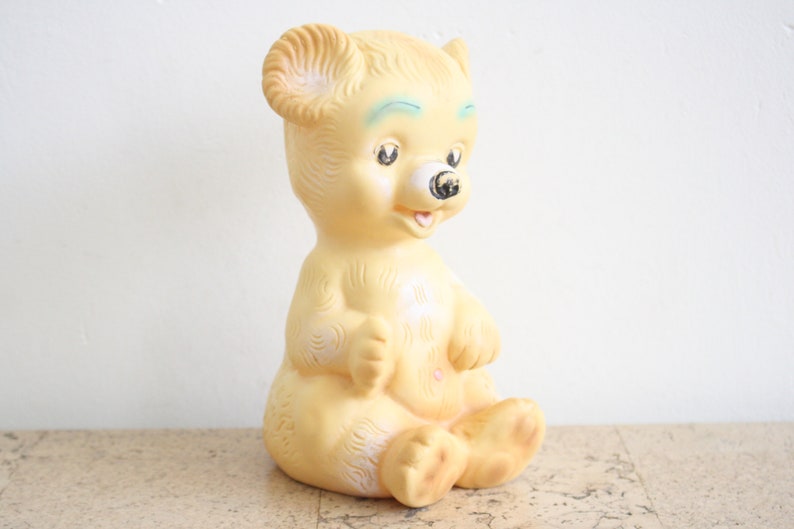 May include: A vintage yellow rubber toy bear with blue eyes and a black nose. The bear is sitting with its legs crossed and has a small smile.