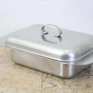 Puede incluir: Una fuente rectangular de acero inoxidable con tapa a juego. La tapa tiene un asa curvada. La fuente tiene un borde ligeramente ensanchado. El metal tiene un acabado cepillado.