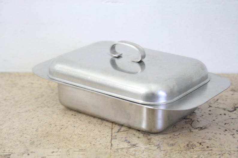 Puede incluir: Una fuente rectangular de acero inoxidable con tapa a juego. La tapa tiene un asa curvada. La fuente tiene un borde ligeramente ensanchado. El metal tiene un acabado cepillado.