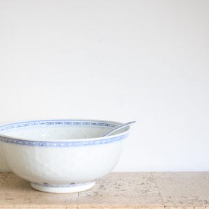 May include: A white porcelain bowl with a blue patterned rim and a matching spoon. The bowl is on a light-coloured surface against a white background. The bowl has a footed base.