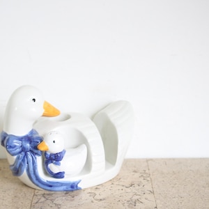 Può includere: Una statuetta di anatra in ceramica bianca con un piccolo anatroccolo accanto. Entrambe le anatre hanno becchi arancioni e sono decorate con fiocchi blu dipinti. L'anatra grande ha un'apertura circolare sul dorso e una finitura lucida.