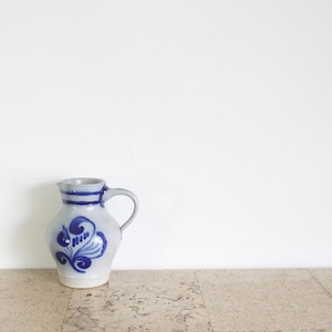 Puede incluir: Una pequeña jarra de cerámica blanquecina con asa y diseño floral azul. La jarra tiene un borde gris y una banda azul alrededor del cuello. El diseño floral es un remolino estilizado. La jarra está sobre una superficie clara.