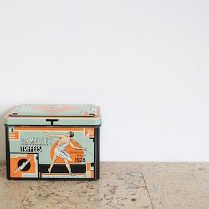 May include: A vintage tin box with a black, orange, and green design. The box features a graphic of a man in a white toga throwing a javelin. The text on the box reads "Van Melle's Toffees Olympic Special 1928".