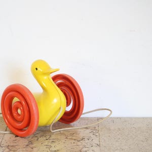 May include: A vintage pull-along toy duck with a yellow body and orange spiral wheels. The duck has a long neck and a beak, and is attached to a white string. A classic toy for children.