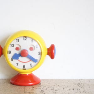 May include: A colourful, vintage-style clock with a yellow face, red base and ear-like accents. The clock face has a smiling face design with a red nose, blue hands, and a white background. A fun, decorative clock.