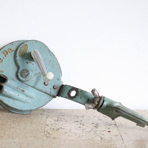 May include: A vintage, light blue metal can opener with a crank handle and a clamp. The can opener has the words "Pe-De" printed on it.