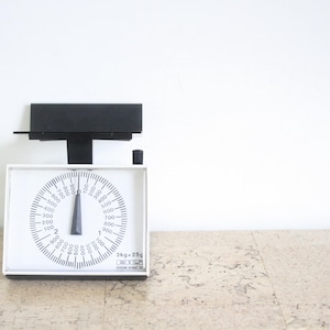 May include: A vintage white and black kitchen scale with a circular dial and a black indicator needle. The scale has a black rectangular top and a base with measurements in grams. The scale is on a light-coloured surface.