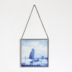 Op de afbeelding: Vierkant ingelijst kunstwerk met een zeilboot op het water, in blauw- en wittinten. Het heeft een ketting om op te hangen. Het kunstwerk is vierkant en hangt aan een witte muur. De stijl doet denken aan aquarel.