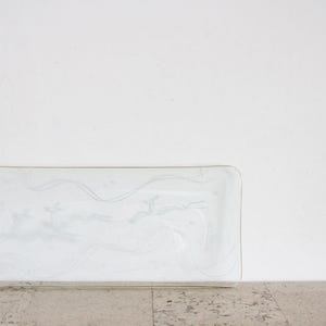 Könnte beinhalten: Ein rechteckiges Serviertablett aus klarem Glas mit abgerundeten Ecken. Das Tablett hat ein zartes geätztes Design aus stilisierten Bäumen und fließenden Linien. Das Tablett wird auf einer hellen Oberfläche vor weißem Hintergrund präsentiert.